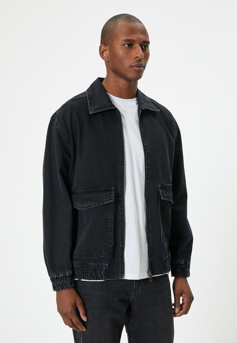 Hombre de pie vestido con una chaqueta negra con cremallera y puños elásticos sobre una camiseta blanca y pantalones negros, frente a un fondo plano.