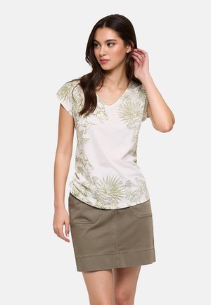 Joven con una camiseta blanca de cuello en V con estampados botánicos verdes y una falda caqui, de pie con una mano cerca de la oreja.