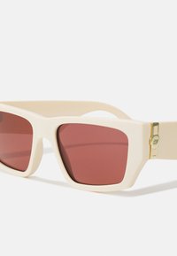 Occhiali da sole color crema con lenti quadrate di tonalità rossa. Accenti di hardware dorato notevoli sui lati della montatura. Finitura matte liscia.