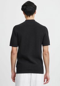 Polo shirt negru cu mânecă scurtă, realizat dintr-un material tricotat texturat, având guler și manșete ribbate, cu o linie dreaptă a tivului și o etichetă de brand subtilă.