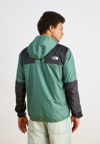 Leichte grüne und schwarze Kapuzenjacke mit elastischen Bündchen und einem Logo auf dem oberen Rücken. Glatte Textur mit kontrastierender Farbgestaltung der Ärmel.