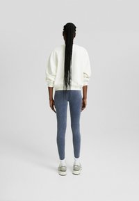 Vit sweatshirt med puffärmar, i kombination med mörkblå leggings. Modellen har på sig vita strumpor och grå sneakers. Håret är stylat i långa flätor.