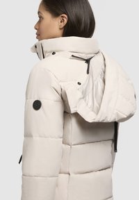 Beige wattierte Jacke mit hohem Kragen, verstellbarem Kapuze, schwarzen Akzenten und Druckknöpfen. Sie hat eine glatte Textur und ein gestepptes Design.