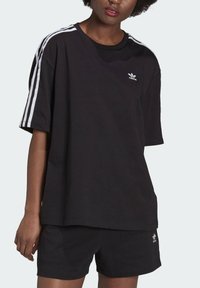 Black oversized t-shirt made of cotton, featuring white three-stripe accents on sleeves and a small white logo on the chest.