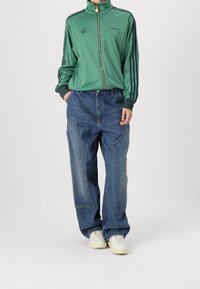 Osoba ubrana w zieloną kurtkę Adidasa na zamek błyskawiczny z czarnymi paskami oraz luźne niebieskie dżinsy z kieszeniami naszywanymi, stojąca z rękami częściowo w kieszeniach.