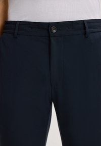 Dunkelblaue Hose aus glattem Stoff, mit Knopfverschluss, Gürtelschlaufen und dezenten Nähten an der Vorder- und Seite.
