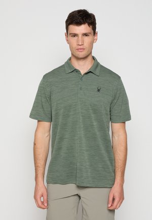 Homme portant un polo à manches courtes vert avec un logo d'araignée noire sur la poitrine, debout devant un fond gris clair uni.