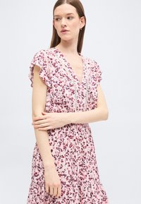 Γυναίκα με ροζ φόρεμα με λουλουδάτο σχέδιο, φαρδιά μανίκια και V-λαιμόκοψη, όρθια με σταυρωμένα τα χέρια, κοιτάζοντας προς τα πλάγια σε απλό φόντο.