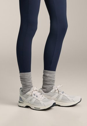 Weiße Sportschuhe mit grauen Akzenten, getragen mit grauen Rippsocken, kombiniert mit marineblauen Leggings. Die Schuhe verfügen über Mesh-Einsätze und Schnürsenkel.