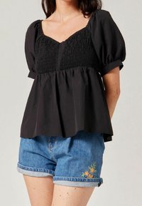 Black short-sleeve blouse with a smocked bodice and loose peplum design, paired with denim shorts featuring floral embroidery on hem.