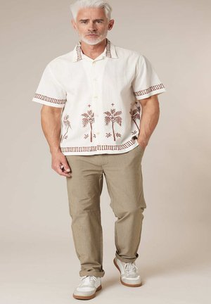 Homme aux cheveux blancs portant une chemise crème à manches courtes avec des imprimés de palmiers, un pantalon beige et des baskets blanches, debout devant un fond uni.