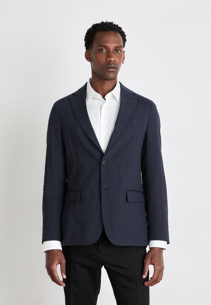 Blazer marine en tissu texturé, à un bouton avec deux poches, associé à une chemise blanche à boutons et un pantalon noir.