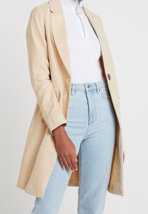 Personne portant un manteau à carreaux beige par-dessus un top blanc zippé et un jean bleu clair taille haute, debout avec une main détendue sur le côté.
