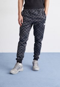 Černé a šedé tepláky s bočními pruhy, elastickým pasem a žebrovanými manžetami. Nošeny s světle šedými sportovními botami.