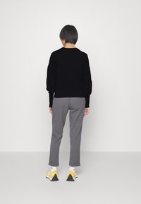 Person med kort hår som bär en svart tröja, grå byxor, vita strumpor och gula och vita sneakers, står vänd mot en enkel vit vägg.