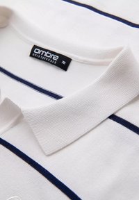 Polo bianco con righe blu navy, tessuto testurizzato, colletto corto e un'etichetta nera con la scritta "ombre" sulla parte interna del colletto.