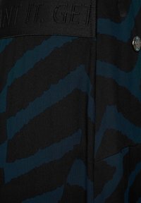 Černá látka s modrým a tyrkysovým abstraktním vzorem. Pas má texturované písmo a stříbrný knoflíkový akcent.