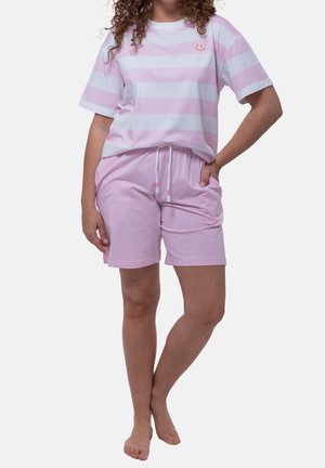 Vrouw die een roze-wit gestreept T-shirt met korte mouwen en effen roze shorts met trekkoord draagt, staat blootsvoets met één hand in de zak.