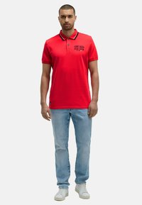 Uomo che indossa una polo rossa con del testo sul petto, jeans chiari e sneaker bianche, in piedi contro uno sfondo neutro.