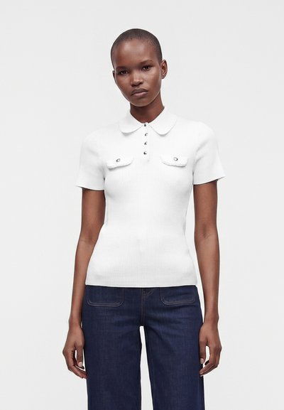 Femme portant un polo blanc à manches courtes côtelé avec des détails de boutons et un jean bleu foncé, debout devant un fond uni.