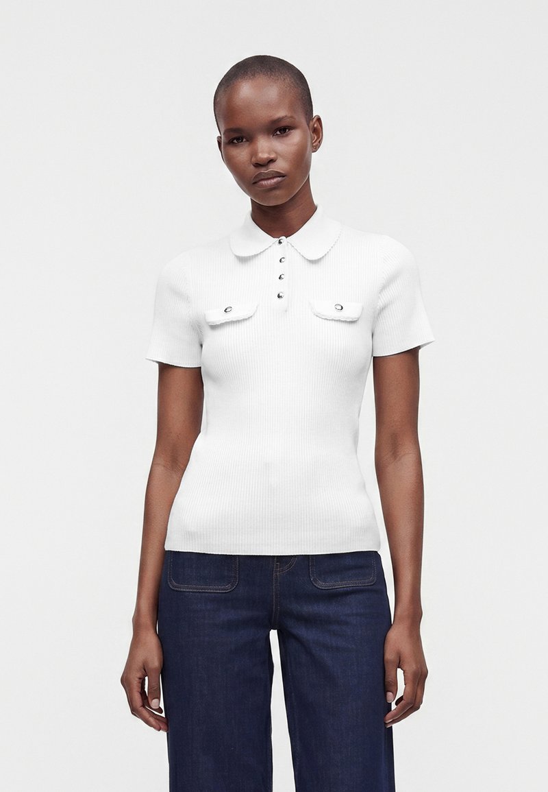 Femme portant un polo blanc à manches courtes côtelé avec des détails de boutons et un jean bleu foncé, debout devant un fond uni.