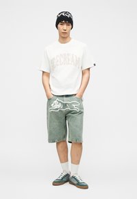 Νεαρός άνδρας που φοράει λευκό μπλουζάκι με την επιγραφή "ICECREAM", τζιν σορτς με σχέδιο καρτούν σκύλου, λευκές κάλτσες, πράσινα αθλητικά παπούτσια και μαύρο σκουφάκι με λευκό μοτίβο.