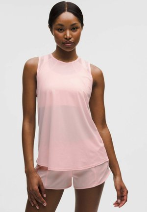 Frau trägt ein hellrosa ärmelloses Oberteil und passende Shorts, steht vor einem einfarbigen Hintergrund.