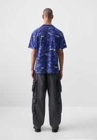 Short-sleeve purple t-shirt with a textured pattern and green accents, paired with baggy dark gray cargo pants featuring multiple pockets.