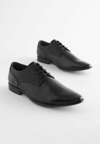 Zapatos de cuero negros con parte superior de tela texturizada, punta afilada y cierre con cordones. Presentan un tacón bajo y paneles contrastantes.