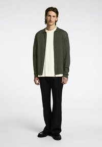 Tamsiai žalia užtraukiama megztinė su apykakle, dėvima virš grietinėlės spalvos marškinėlių, derinama su laisvomis juodomis kelnėmis ir juodais batais.