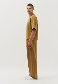 Mosterdgele top met korte mouwen en bijpassende broek, gemaakt van gladde stof. Model draagt witte sneakers en staat naar links gekeerd tegen een effen achtergrond.