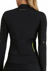 Veste de sport noire à manches longues, avec des panneaux en maille texturée, un logo vert réfléchissant et des accents, ainsi qu'une poche zippée sur le côté pour le rangement.