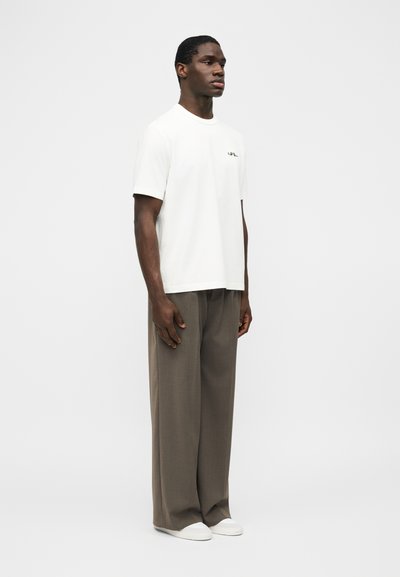 Άνδρας που στέκεται φορώντας λευκό μπλουζάκι με κοντά μανίκια, φαρδιά καφέ παντελόνια και λευκά αθλητικά παπούτσια σε απλό φωτεινό φόντο.