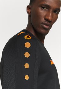 Maglietta sportiva nera con accenti arancioni, incluso un logo e un motivo a pois sulla spalla. Tessuto testurizzato con design aderente.