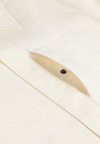 Cămașă bej deschis, cu un buzunar pe piept prevăzut cu închidere cu nasture, fabricată dintr-un material țesut cu o textură fină și detalii de cusătură vizibile.