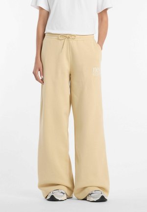 Person iført beige vidde sweatpants med snøre i taljen og hvide sneakers, stående mod hvid baggrund.