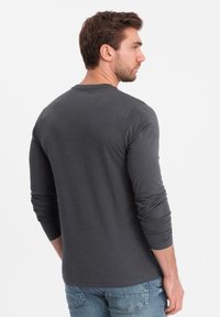 Ein Mann trägt ein dunkelgraues Langarmhemd und blauwe Jeans. Er steht seitlich mit dem Kopf nach rechts gedreht vor einem weißen Hintergrund.