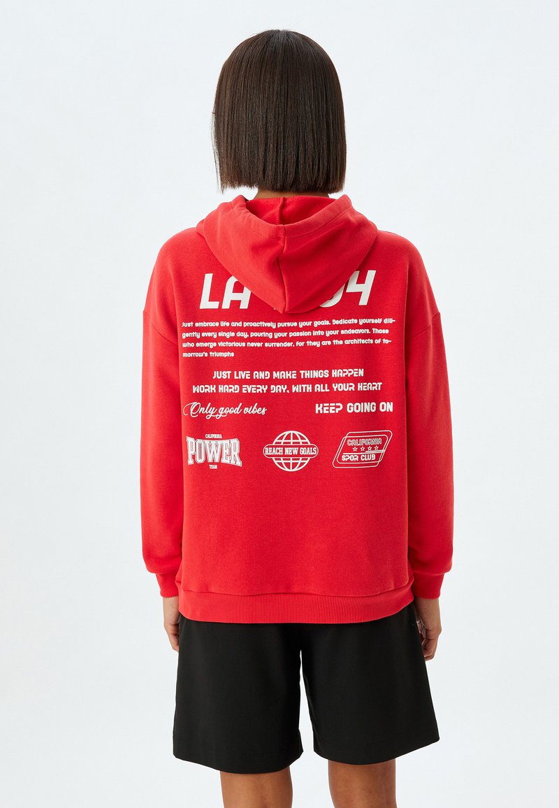 Röd huvtröja med vit text och grafik på baksidan, med avslappnad passform och ribbade muddarna. Bärs med svarta shorts.