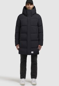 Piumino blu navy con colletto alto e cappuccio regolabile. Presenta una chiusura con zip, pannelli cuciti e una patch con logo sull'orlo inferiore.