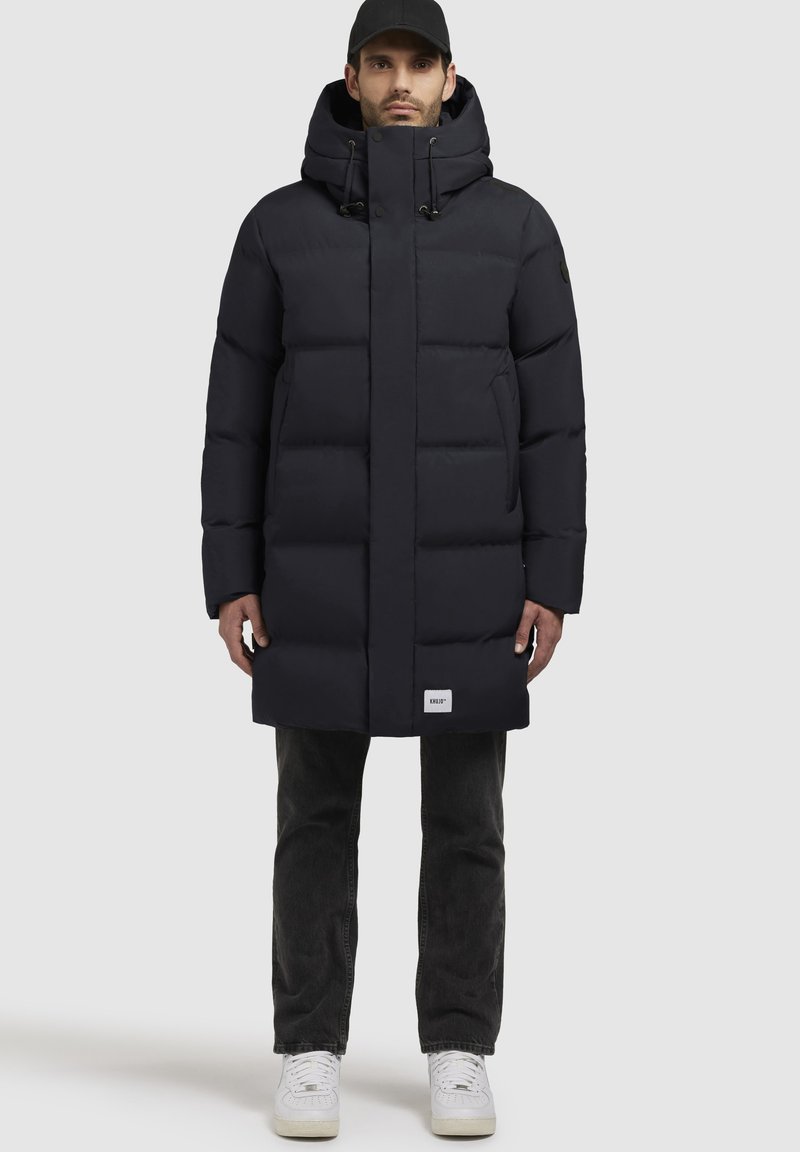 Piumino blu navy con colletto alto e cappuccio regolabile. Presenta una chiusura con zip, pannelli cuciti e una patch con logo sull'orlo inferiore.