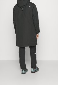 Abrigo largo negro impermeable con capucha, que cuenta con un cierre de lazada y detalle de logo. Combinado con pantalones negros y zapatillas deportivas gris-azules.