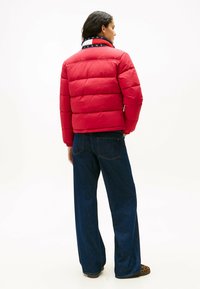 Rød pufferjakke med quiltet design og elastiske manchetter, parret med mørkeblå brede jeans. Jakken har en brandet krave.