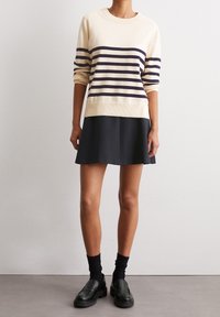 Maglione crema con righe navy, polsini e orlo a coste, abbinato a una gonna nera a sbalzo. Mocassini neri e calzini alla caviglia completano il look.