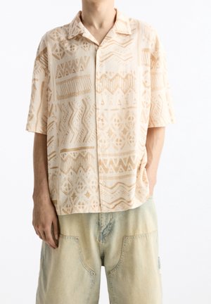 Homme portant une chemise beige à manches courtes avec des motifs abstraits et un pantalon en denim bleu clair ample, sur fond blanc.