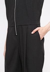 Vêtement noir avec fermeture éclair à l'avant et poche latérale, main insérée dans la poche, vue en gros plan de la taille et des hanches.