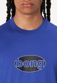 Mėlyna medvilnės marškinėlių su apvaliu juodu logotipu, ant kurio baltais rašmenimis užrašyta "bong" per krūtinę. Standartinio kirpimo su apvaliu kaklu.