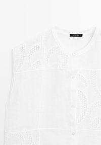 Blouse blanche sans manches avec encolure ronde, boutonnée sur le devant et motifs délicats en dentelle découpée sur la partie supérieure de la poitrine et les épaules.
