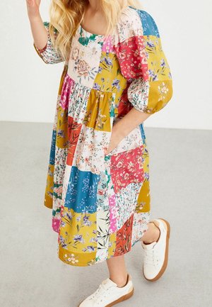 Blomster patchwork kjole med firkantet halsudskæring, puffede ærmer og lommer. Farverne inkluderer gul, pink, blå og hvid med forskellige mønstre.