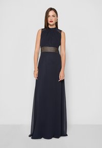 Námornícky modré večerné šaty s bezrukávovým dizajnom, zhromaždeným výstrihom a tkanými kovovými detailmi v oblasti pásu. Plynulo padajúca sukňa po zem.