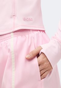 Κοντινό ενός χεριού στην τσέπη ροζ παντελονιού με ελαστική μέση και ροζ μπλούζας με φερμουάρ και το λογότυπο "BORG".